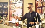 Photo of a bartender wearing a waistcoat and glasses smiling while preparing a cocktail on the bar