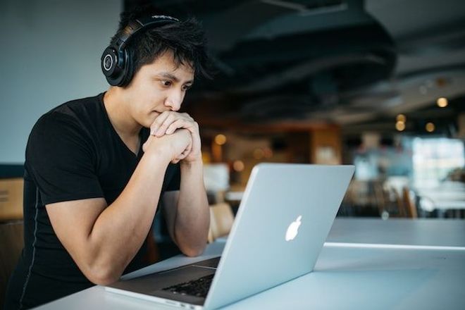 Man wearing headphones, staring at an Apple laptop, and looking slightly anxious