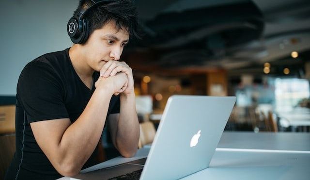 Man wearing headphones, staring at an Apple laptop, and looking slightly anxious