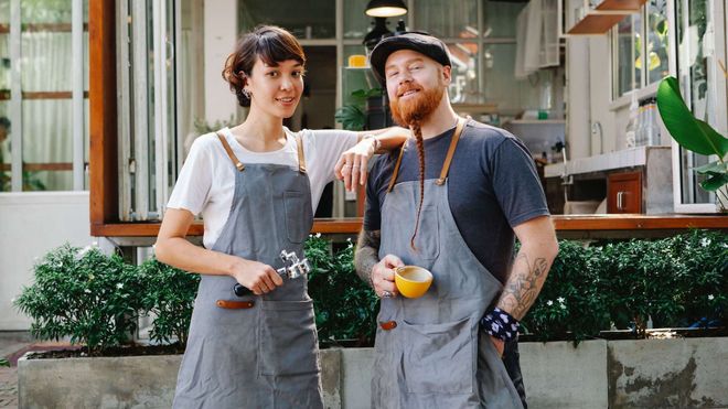 Female and male barista facing the camera and smiling in front of a shop