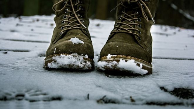 Close up of brown/green winter lace-up boots with snow on them, standing on snowy ground.