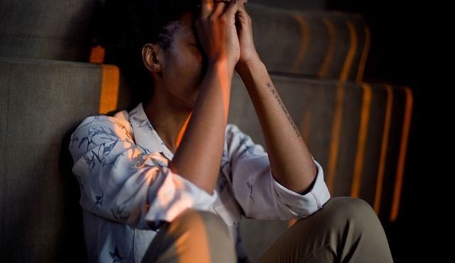Woman leaning against wall, with hands clasped in front of her face and elbows rested on knees