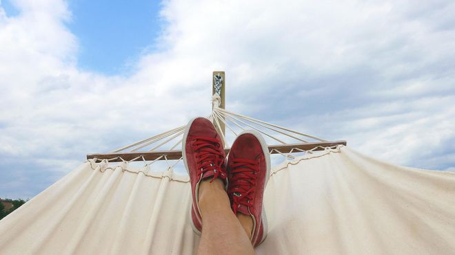 A pair of feet wearing red deck shoes in a canvas hammock with blue sky in the background.