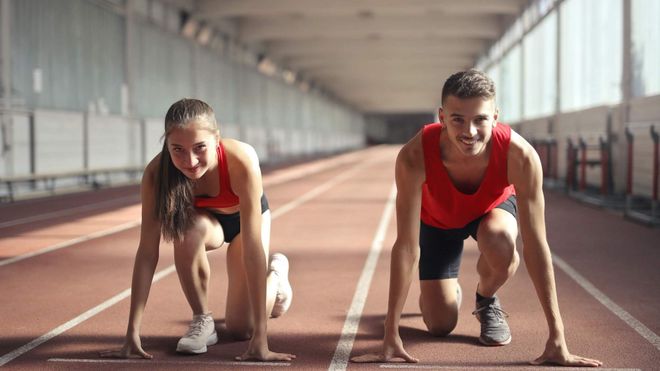 Female and male runner wearing red tank tops crouched on an indoor running track.