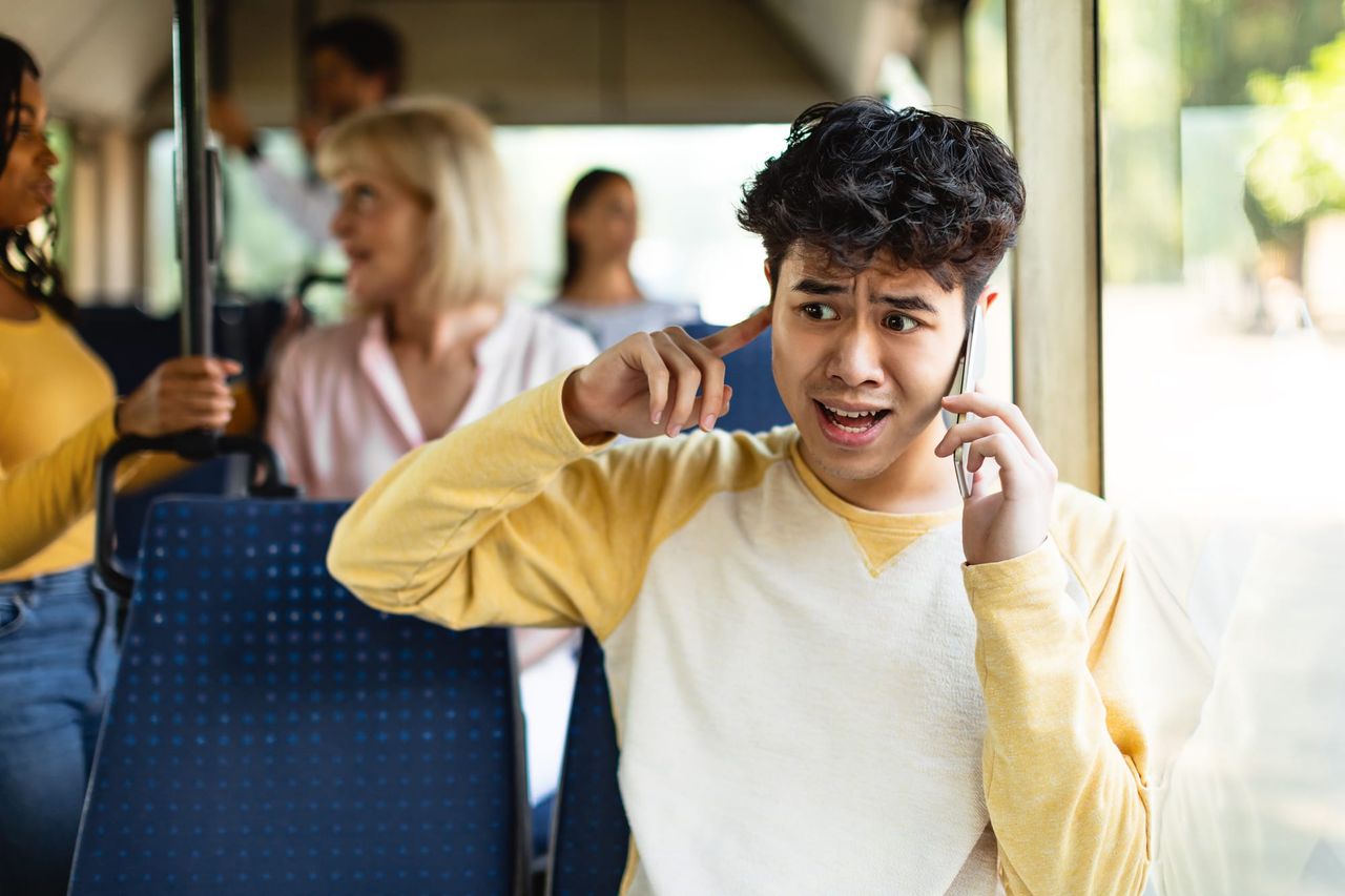 A young man sits on a bus, mobile phone to one ear and a finger in the other, looking frantic.