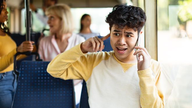 A young man sits on a bus, mobile phone to one ear and a finger in the other, looking frantic.
