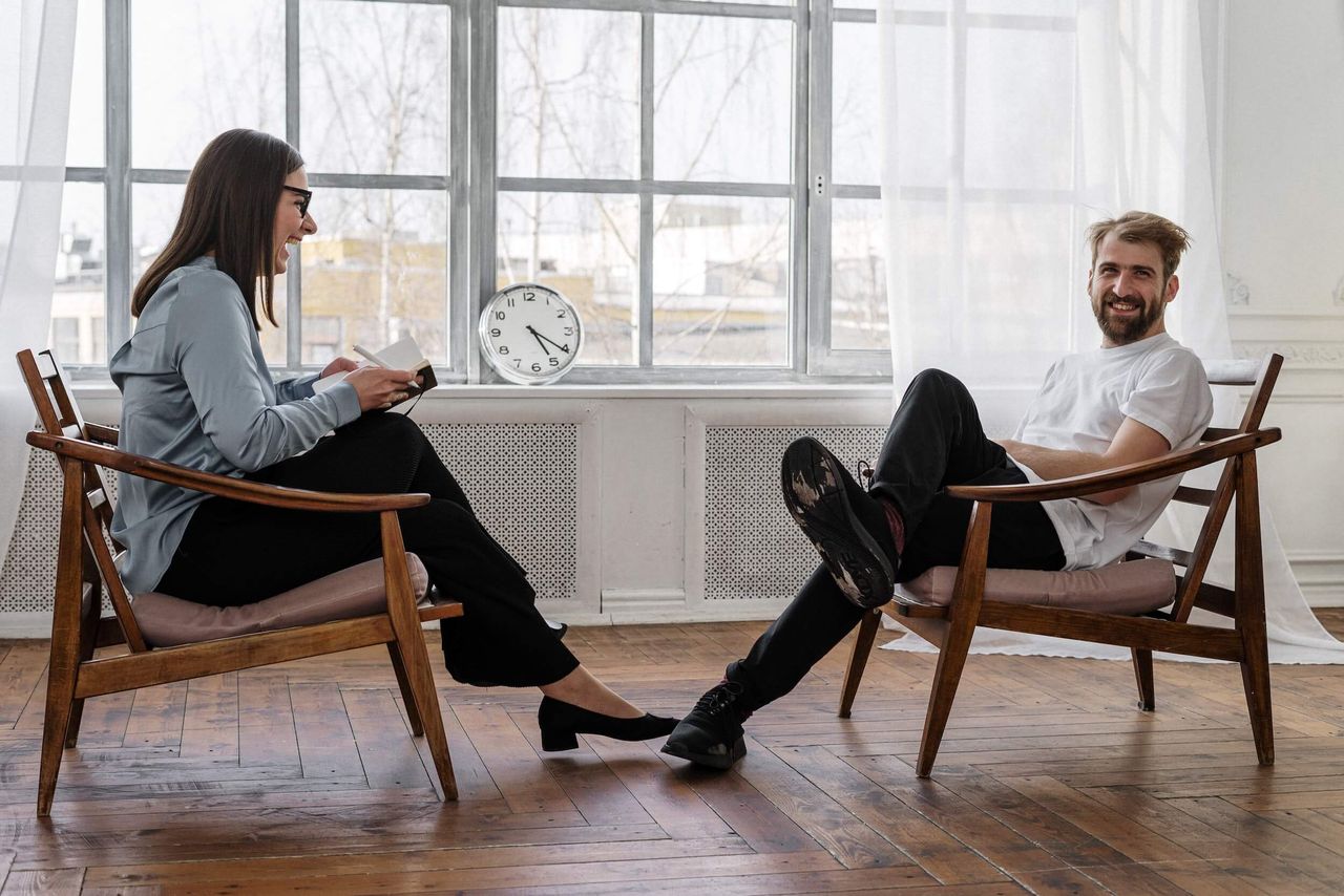 A woman and a man sitting opposite each other on low wooden armchairs, laughing. The man is looking to camera.