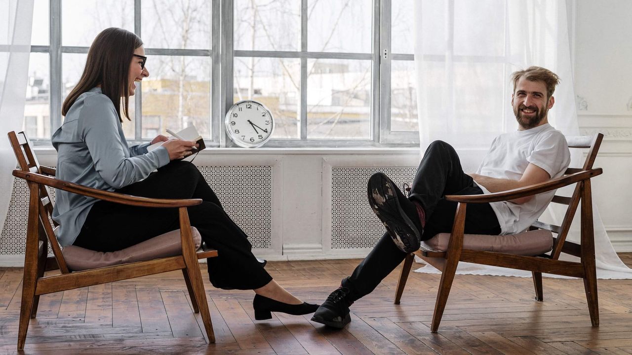 A woman and a man sitting opposite each other on low wooden armchairs, laughing. The man is looking to camera.