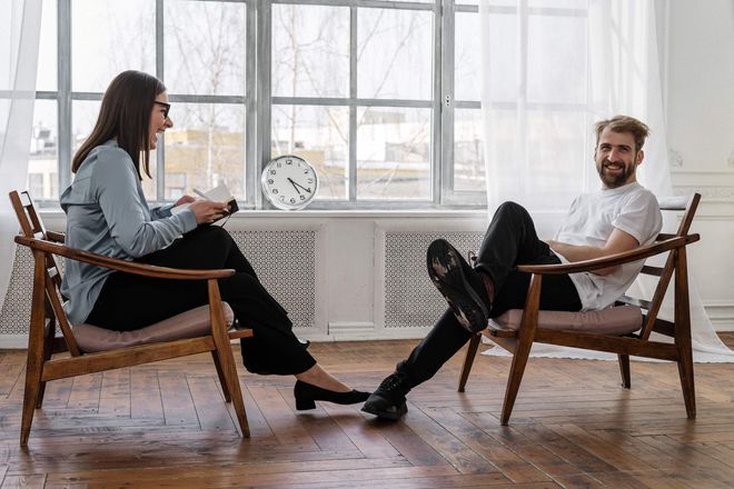 A woman and a man sitting opposite each other on low wooden armchairs, laughing. The man is looking to camera.