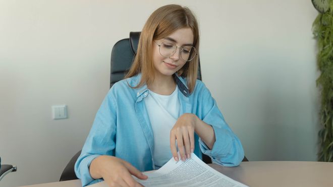 Woman wearing glasses and a light blue shift smiling while reading a paper document.