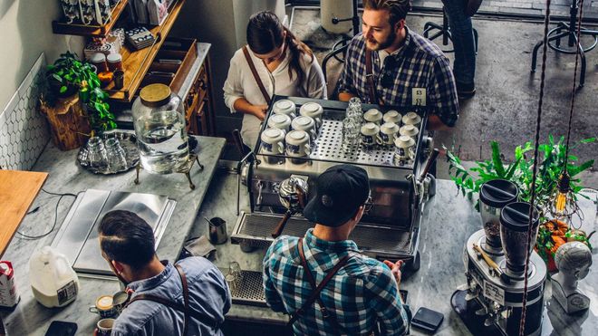A pair of baristas preparing coffee in a coffee shop while a man and woman wait on the other side of the counter.