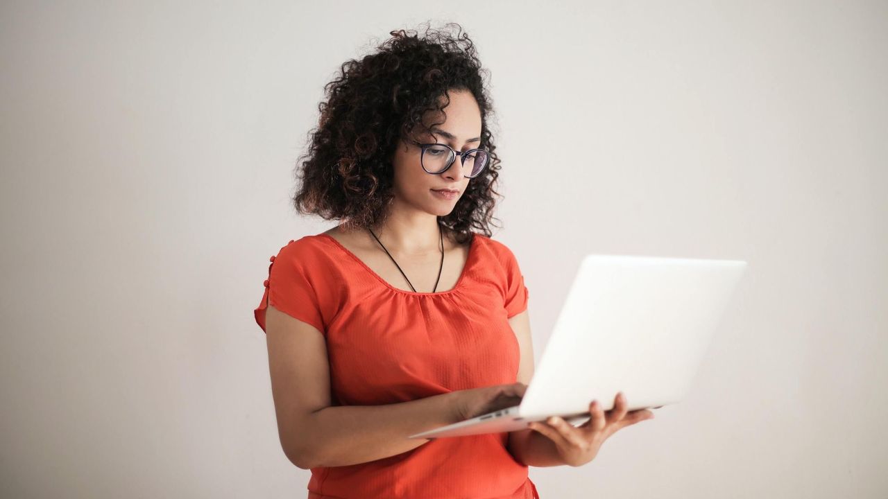 Woman with dark, curly hair and glasses holding a silver laptop computer standing in front of a white background.
