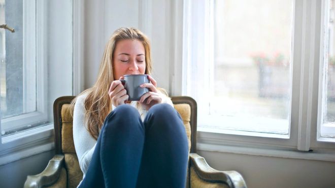Photo of a woman sitting in an armchair with her knees up, sipping from a ceramic mug