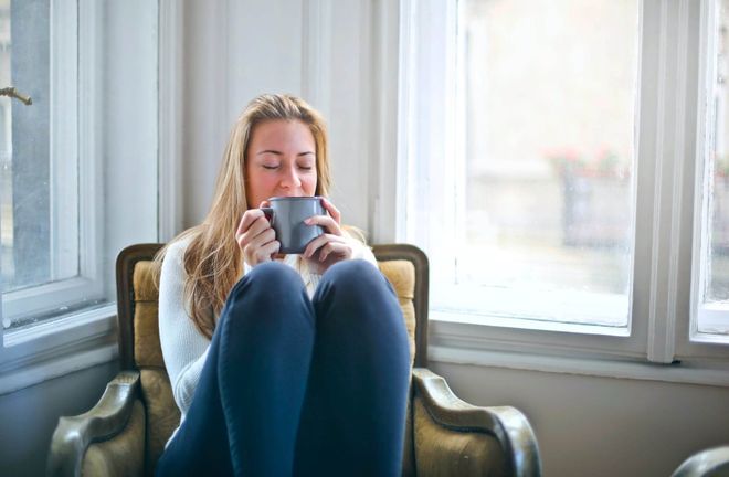Photo of a woman sitting in an armchair with her knees up, sipping from a ceramic mug
