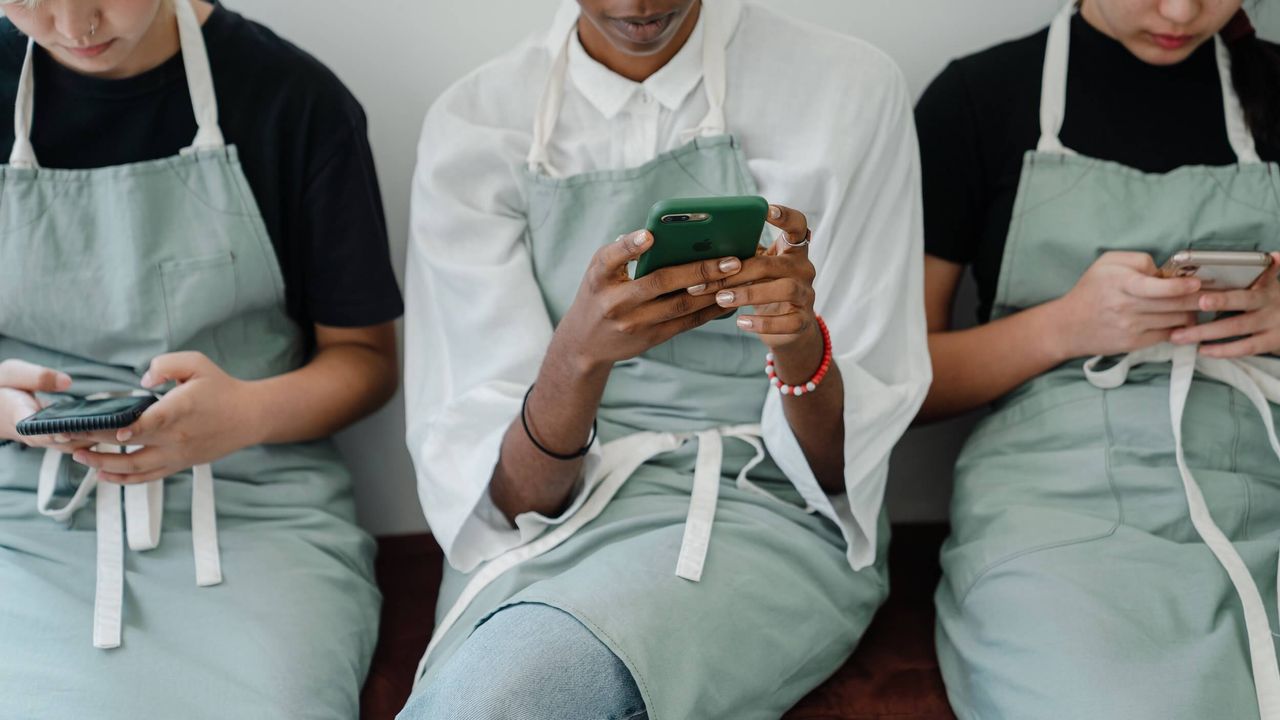 Photo of three staff all wearing aprons, seated in a row and looking at their phones 