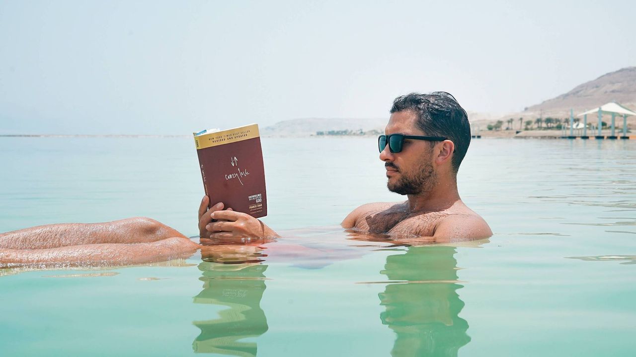 Man wearing sunglasses floating in the sea reading a book.