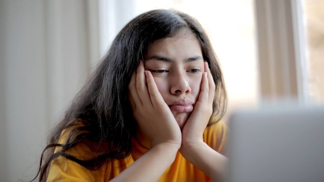 Young woman with dark hair looking bored while resting her head in her hands and looking at a laptop.