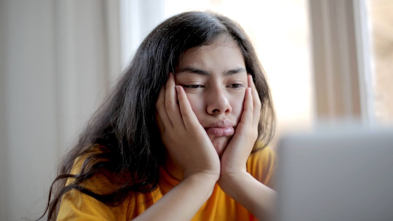 Young woman with dark hair looking bored while resting her head in her hands and looking at a laptop.