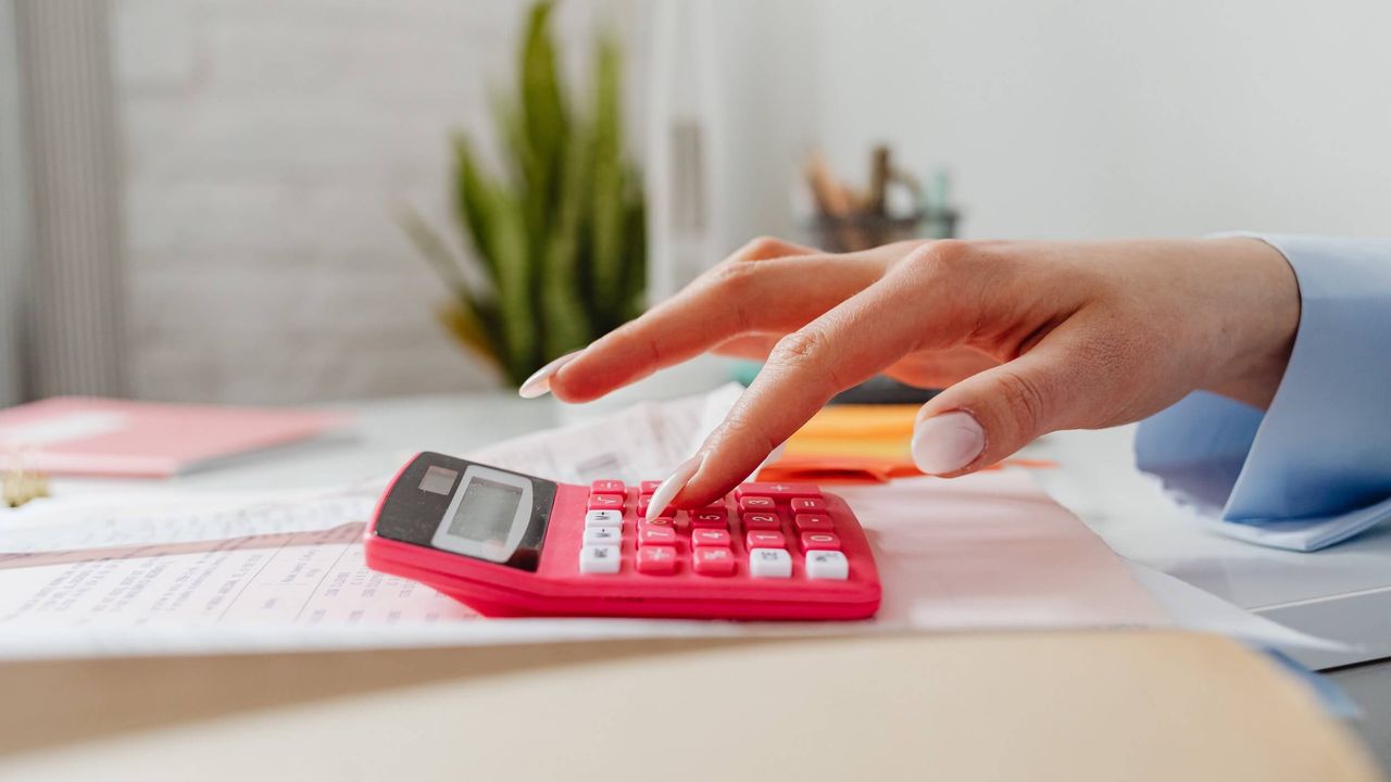 Photo of woman's hand using a light red calculator resting on a desk.