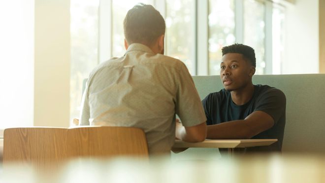 Two men sat opposite each other at a canteen-like booth, one man looking worried