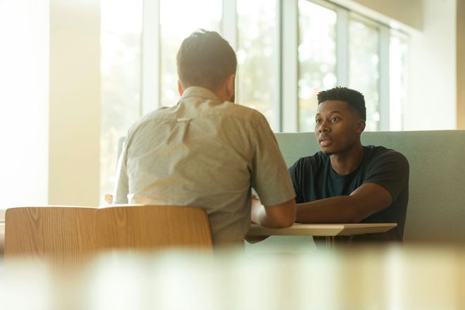 Two men sat opposite each other at a canteen-like booth, one man looking worried
