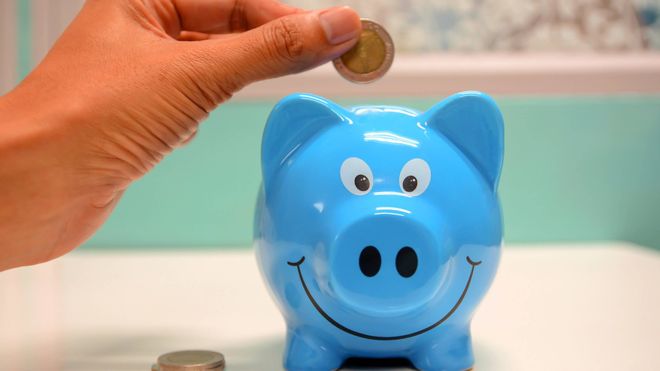 Photo of a hand placing a coin into a blue piggy bank with a smiling face