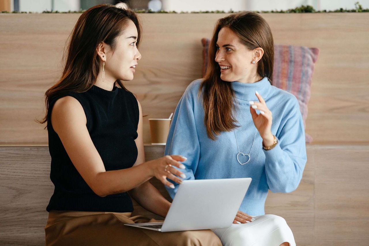 Two young professional women in animated conversation. The woman on the left has a laptop computer on her lap.