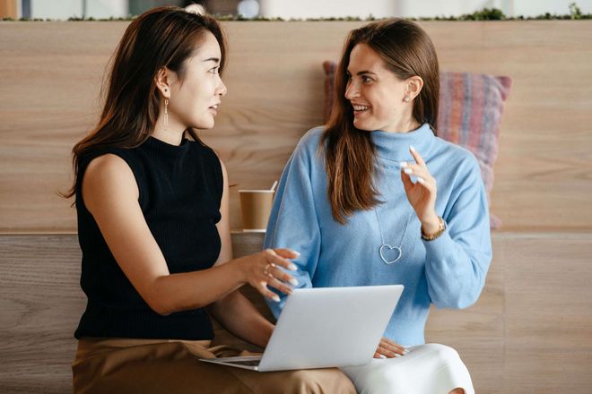 Two young professional women in animated conversation. The woman on the left has a laptop computer on her lap.