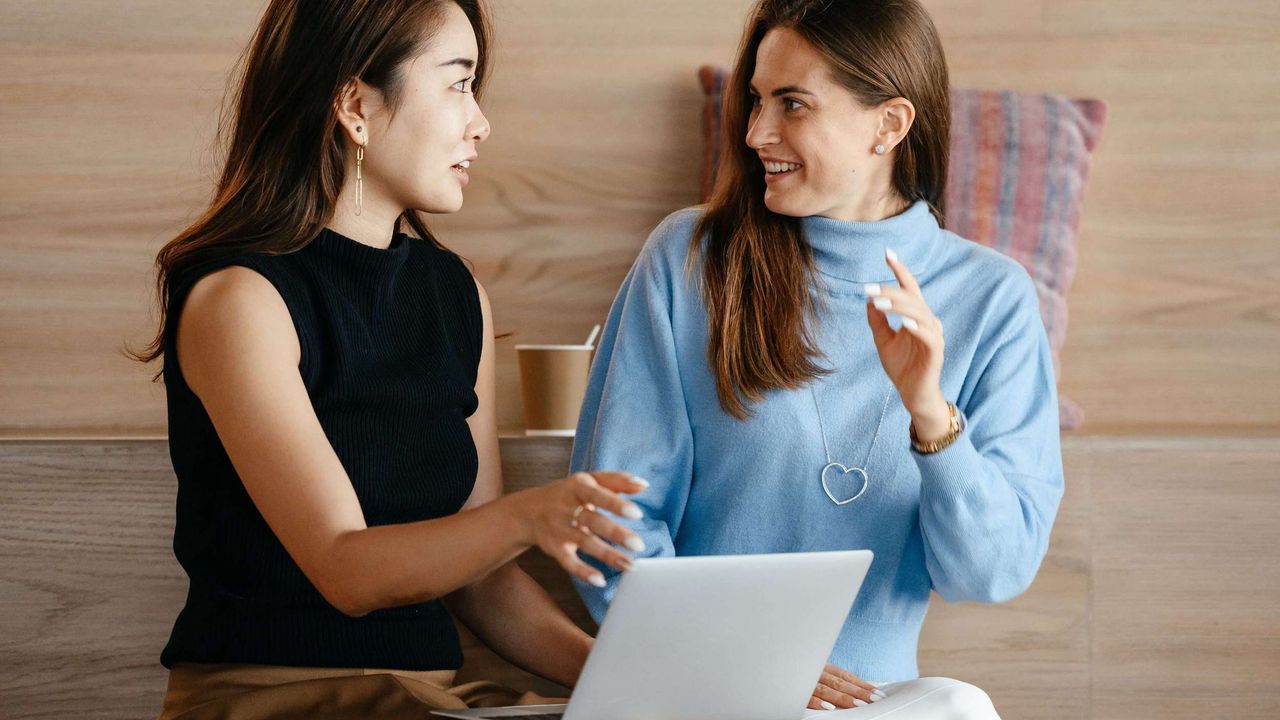 Two young professional women in animated conversation. The woman on the left has a laptop computer on her lap.