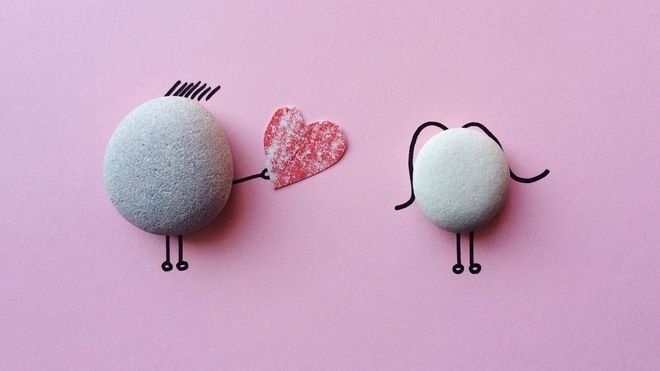 Two grey pebbles on pink paper with doodles of one handing a heart to the other.