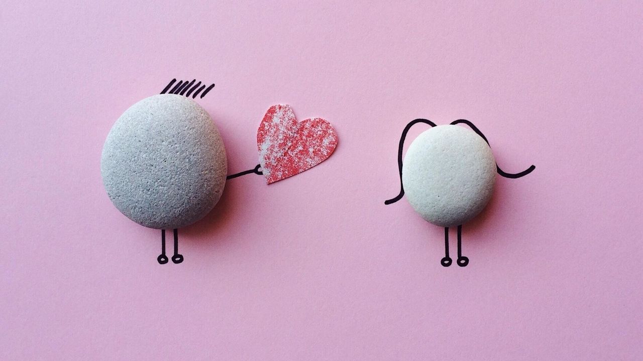 Two grey pebbles on pink paper with doodles of one handing a heart to the other.