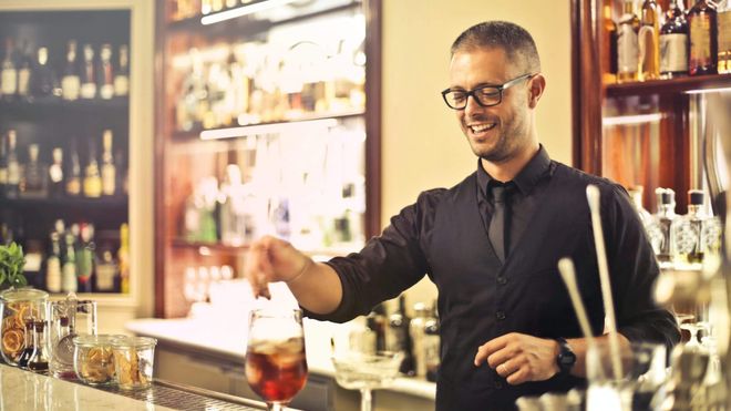 Photo of a bartender wearing a waistcoat and glasses smiling while preparing a cocktail on the bar