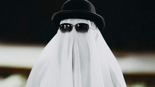 Someone covered in a white sheet to look like a ghost, with sunglasses and a bowler hat over the sheet.