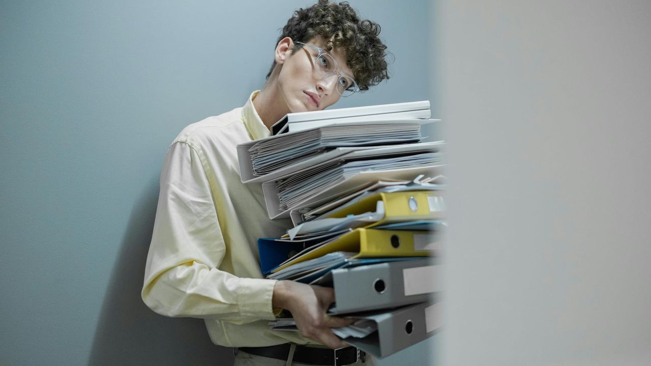 A man in office attire with glasses holds a large pile of files and paperwork, leaning his chin on the top in disdain.