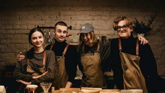 A group of four baristas smile and pose together behind their rustic-style coffee shop counter.