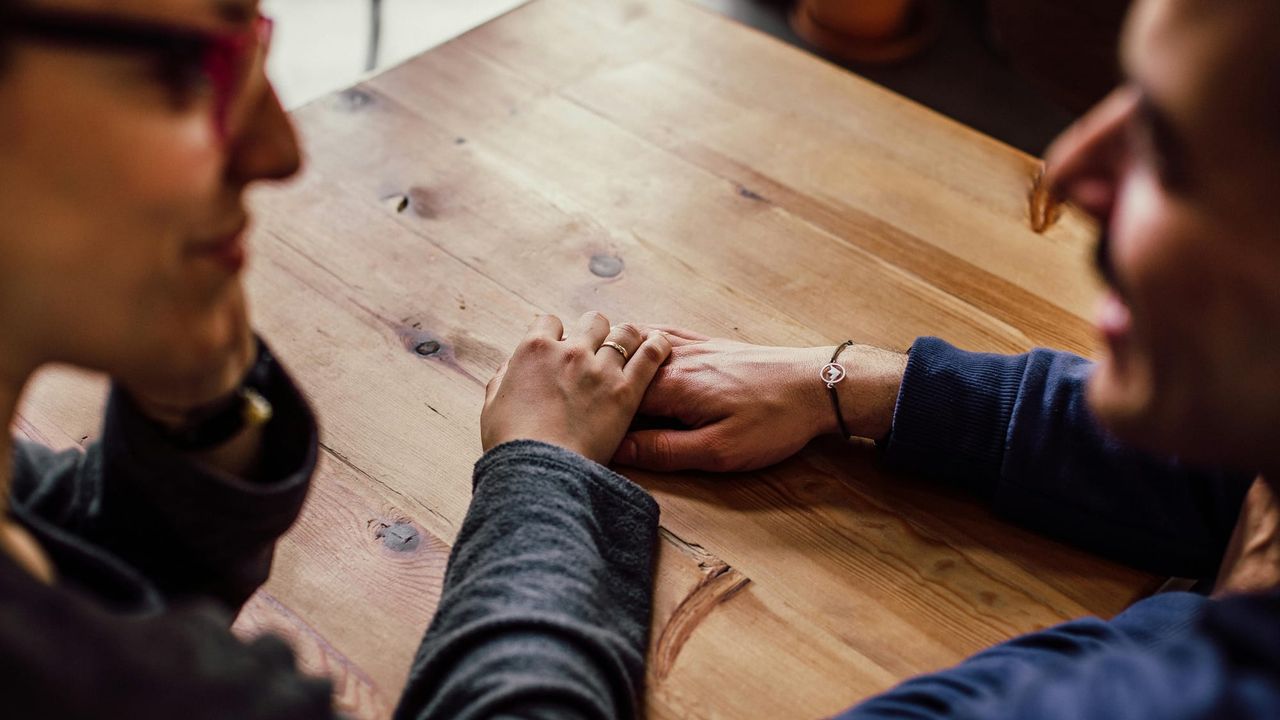 A couple look at each other, sat at a table, while we focus on their hands touching on the tabletop.
