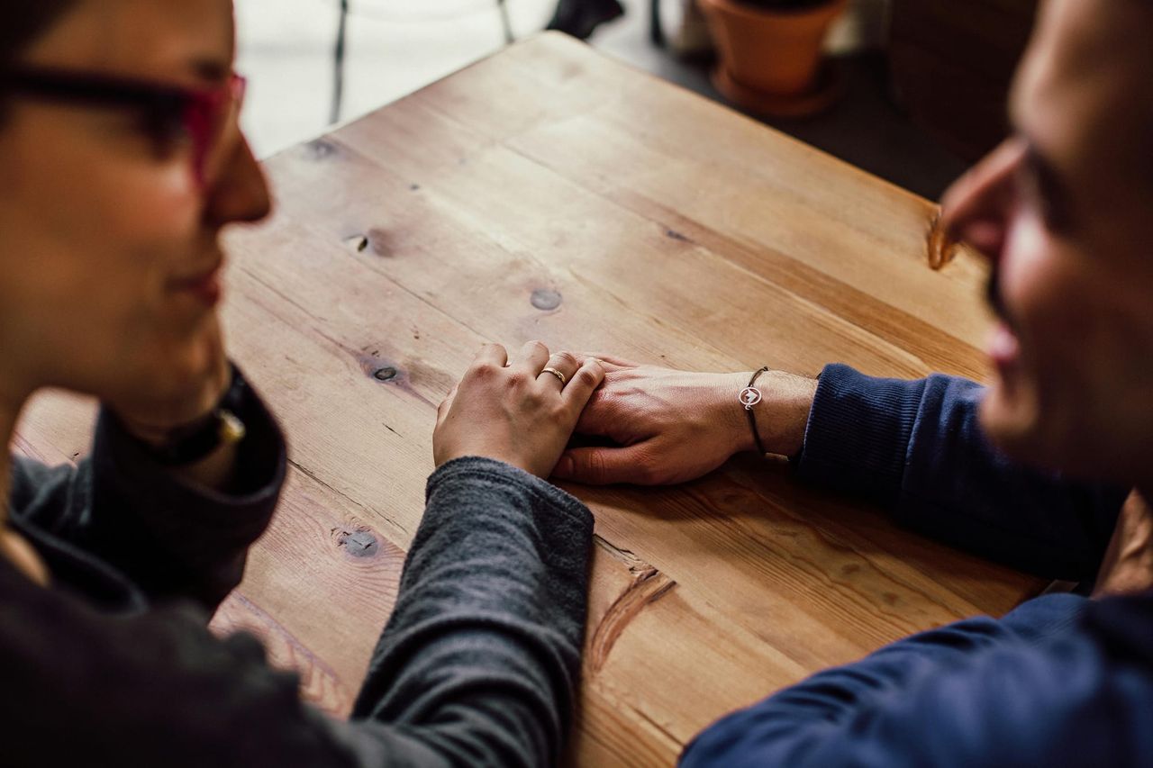 A couple look at each other, sat at a table, while we focus on their hands touching on the tabletop.