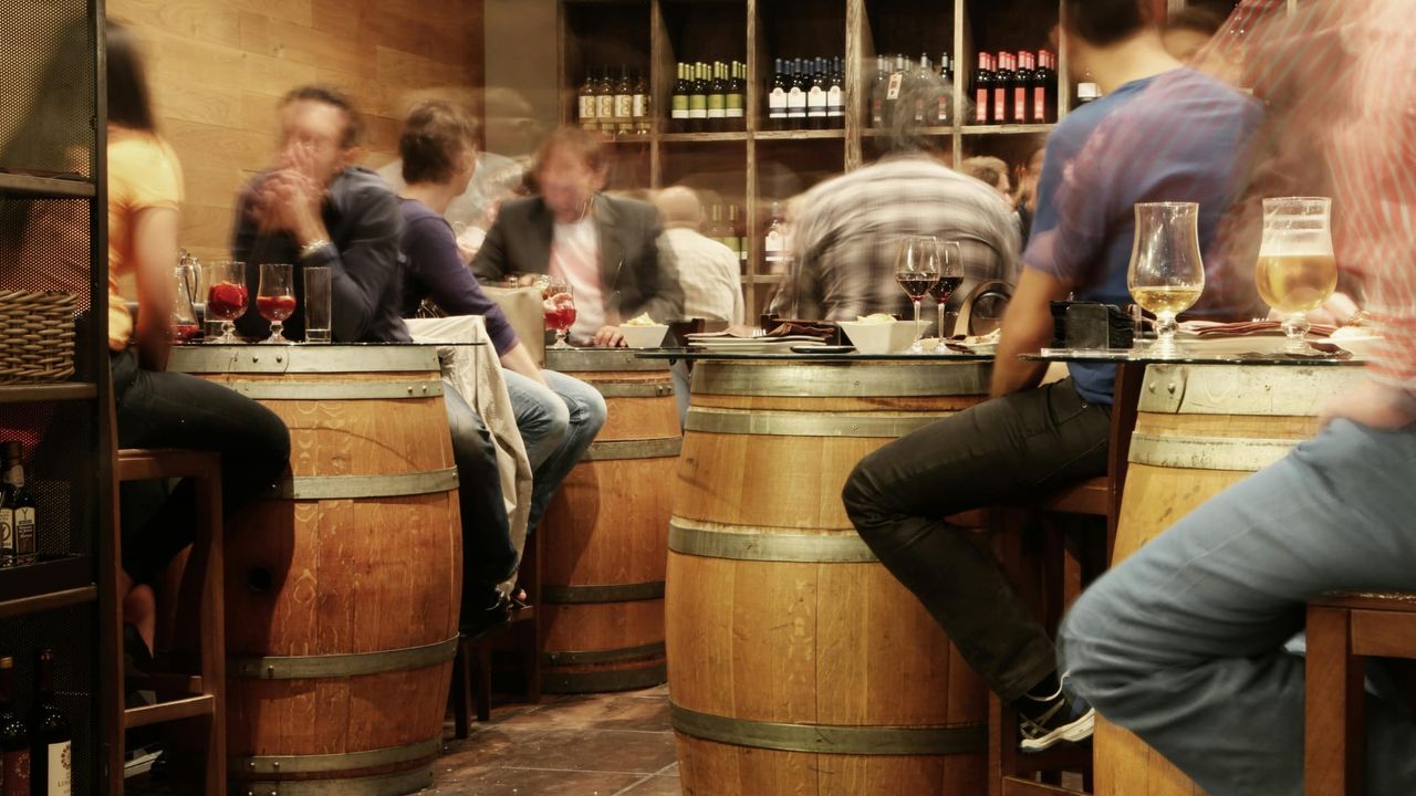 A busy wine bar and brewery, people blurred around barrel-styled tables to suggest a fast-paced setting or  passing of time.