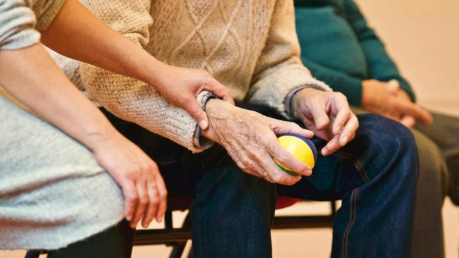 A care worker sat beside an elderly patient, touching their wrist in comfort and support