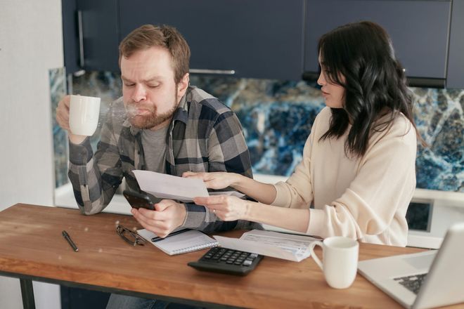 A man spitting out tea in shock as he looks upon a bill a woman shows him