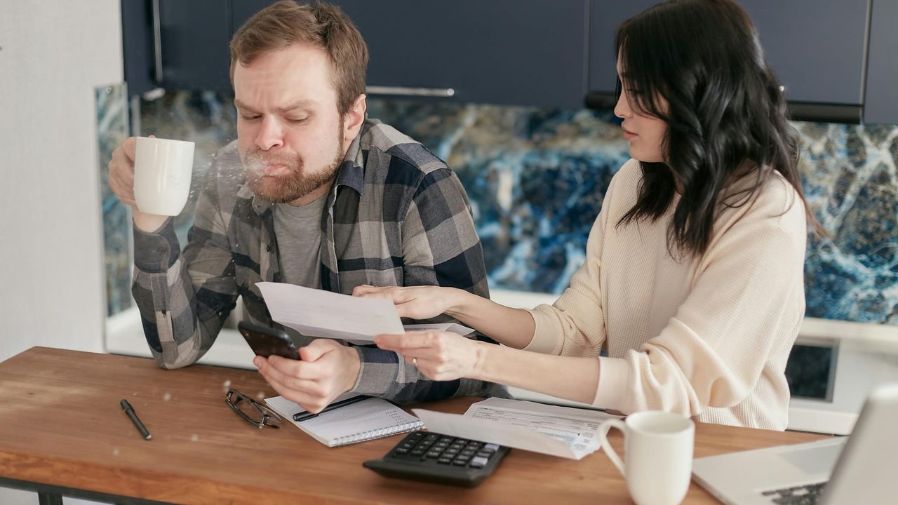 A man spitting out tea in shock as he looks upon a bill a woman shows him