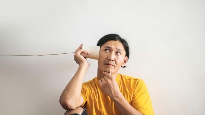 A man in a yellow t-shirt, sat against a white wall with a paper cup to his ear. A string from the cup leads away while he holds his chin, seemingly listening.
