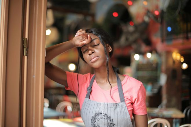 A young female hospitality worker, leaning against a doorframe with her eyes closed and forehead pressed against the back of her hand.
