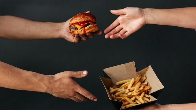 Two people exchanging a burger and a box of chips between them.
