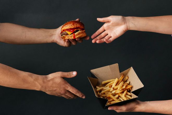 Two people exchanging a burger and a box of chips between them.