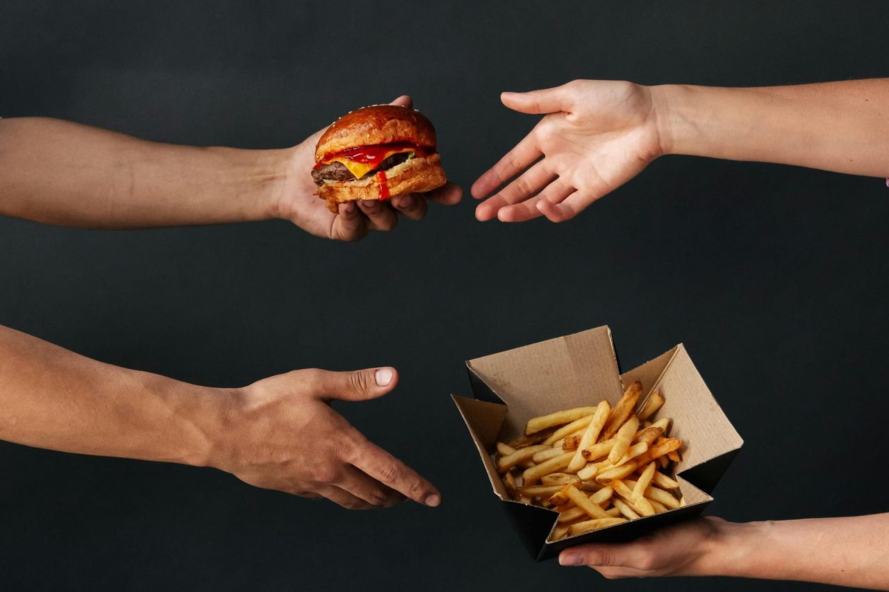 Two people exchanging a burger and a box of chips between them.
