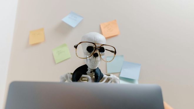 A skeleton wearing glasses and a loose tie sits in front of an open laptop with many Post-it notes plastering the wall behind it.