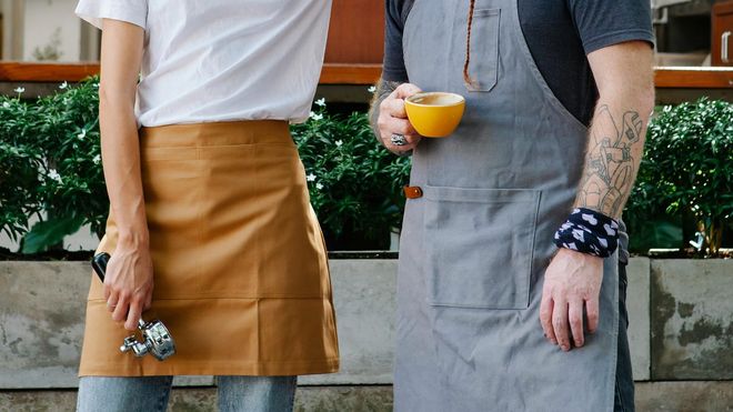 Two baristas from the chest down, both wearing aprons. One holds a yellow coffee cup while the other holds a portafilter. A small green hedge lines behind them.