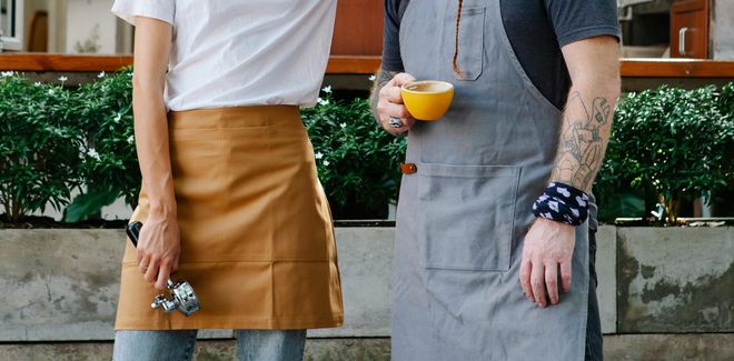 Two baristas from the chest down, both wearing aprons. One holds a yellow coffee cup while the other holds a portafilter. A small green hedge lines behind them.