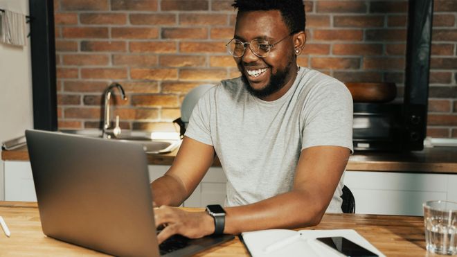 A young man sat at a kitchen counter with an open notebook and a laptop, smiling at the screen as he types.